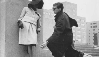 A man kneels before a mannequin wearing a vintage coat and gloves, urban backdrop.